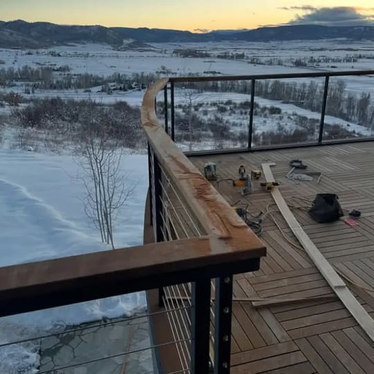 Curved wood-capped cable railing on a parquet-pattern deck overlooking a snowy valley at sunrise
