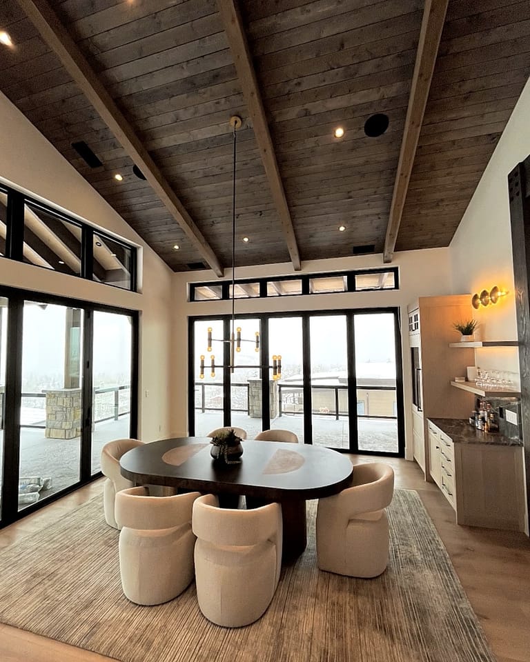 Dining room with an oval dark wood table and cream chairs under a vaulted wood-plank ceiling, with glass doors to a snowy deck