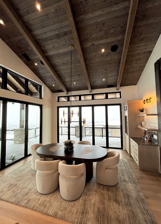 Dining room with an oval dark wood table and cream upholstered chairs under a vaulted wood-plank ceiling, with black-framed glass doors to a snow-covered outdoor space with a railing, and a bar cabinet with a dark counter