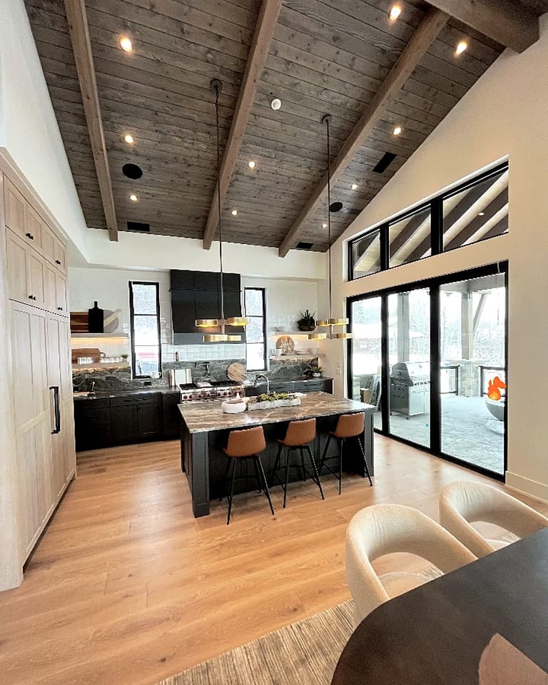 Kitchen with black base cabinets, light-wood tall cabinets, an island with three tan stools and a vaulted wood-plank ceiling
