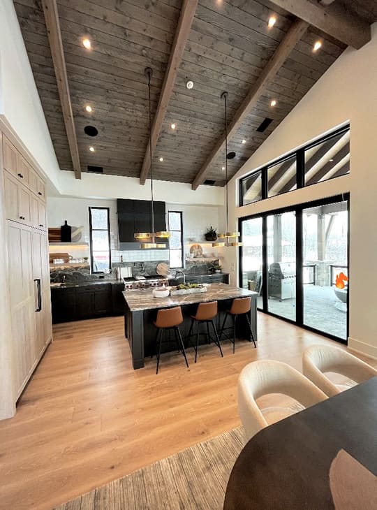 Kitchen with black base cabinets and a wall of light-wood tall cabinets, an island with a veined gray-brown top and three tan upholstered stools, gold-toned pendant lights and a vaulted wood-plank ceiling, opening through black-framed glass doors to a covered outdoor area