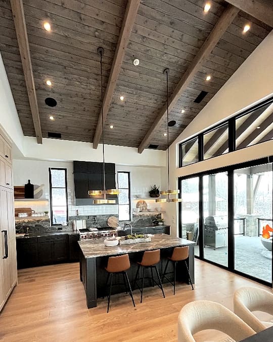 Kitchen with black base cabinets and a wall of light-wood tall cabinets, an island with a veined gray-brown top and tan upholstered stools, and a vaulted wood-plank ceiling