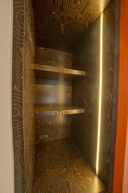 Closet niche lined with patterned plywood and lit by a vertical LED strip